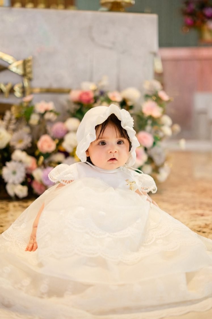 bebé con vestido de bautizo y tocado frente a flores en la iglesia